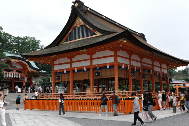 朱色の柵と屋根が特徴的な日本の神社(稲荷神社)の建物と、多くの参拝客が行き交う様子