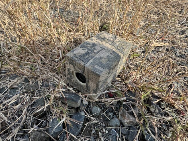 「見えない刃：福岡・博多で急増する位置情報アプリを悪用したデジタル・ストーカー枯れた草と小石の地面に隠された、カモフラージュ柄のハウジングに入った小型監視カメラ。レンズ部分が正面を向いている。対策」