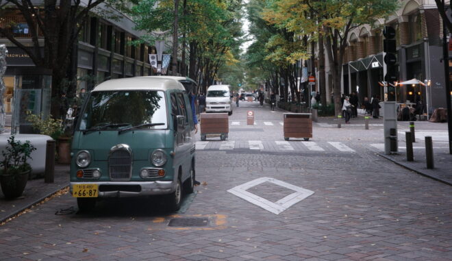 並木道が続く都会の石畳の通り。手前に水色のレトロなバンが駐車され、秋の始まりを感じさせる