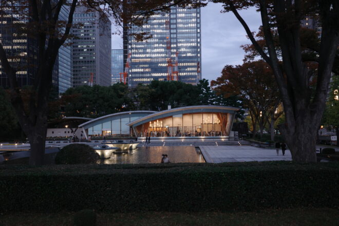 夕暮れ時、都会の高層ビル群を背景にした公園内のモダンなガラス張りのカフェ。カフェ内部は明るく人々で賑わい、前景には水面と暗い植え込みがある。