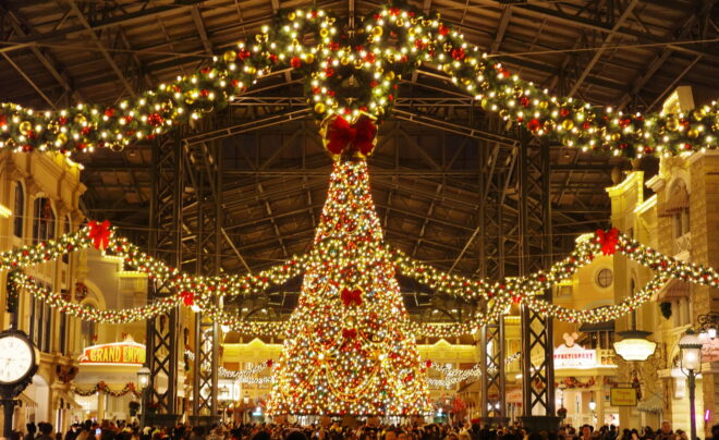 華やかな電飾で飾られた巨大なクリスマスツリーが中央に立つ、屋内アーケード（ワールドバザールのような場所）の夜景。鉄骨構造の天井の下、冬服を着た大勢の訪問者で賑わっている様子。