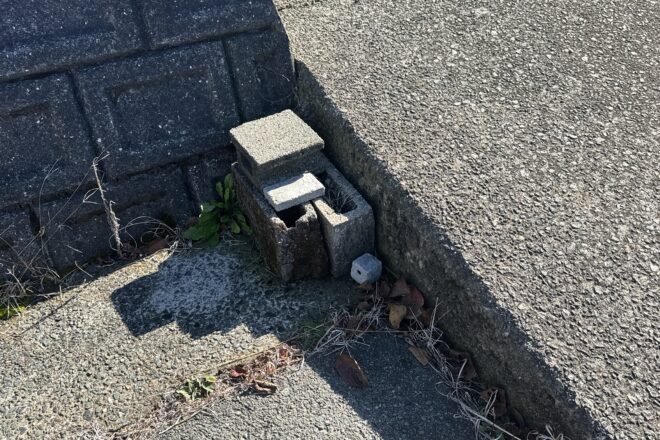 強い日差しの下、アスファルトの路面とコンクリート擁壁の隙間に不自然に積み重ねられた複数の古いコンクリートブロック、および周囲の枯れ草や雑草。