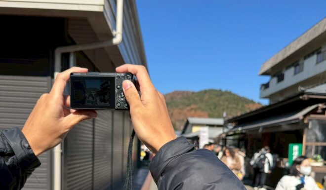 黒いダウンジャケットを着た人物が、コンパクトカメラを両手で構え、賑わう観光地の街並みを撮影している様子。快晴の青空の下、遠景には山並みが見える。