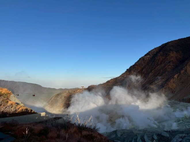 澄み渡る青空の下、山肌から勢いよく白い噴煙が立ち上る大涌谷の風景と、遠くに見えるロープウェイ。