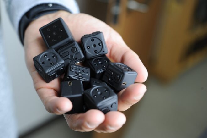 大人の男性の手のひらに大量に載せられた、黒くて小さなサイコロ型の超小型隠しカメラ。赤外線LEDライトやピンホールレンズ、充電用ポートがついている。