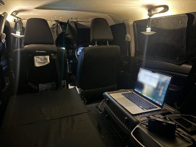 窓が遮光サンシェードで覆われた乗用車の車内。薄暗い照明の中、後部スペースに開いたノートパソコン、カメラ、黒いマットが設置された張り込みや車中泊を連想させる様子。